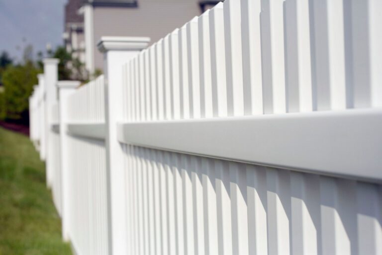 Angled close-up of a white vinyl picket fence with staggered picket tops and decorative post caps beside a green lawn