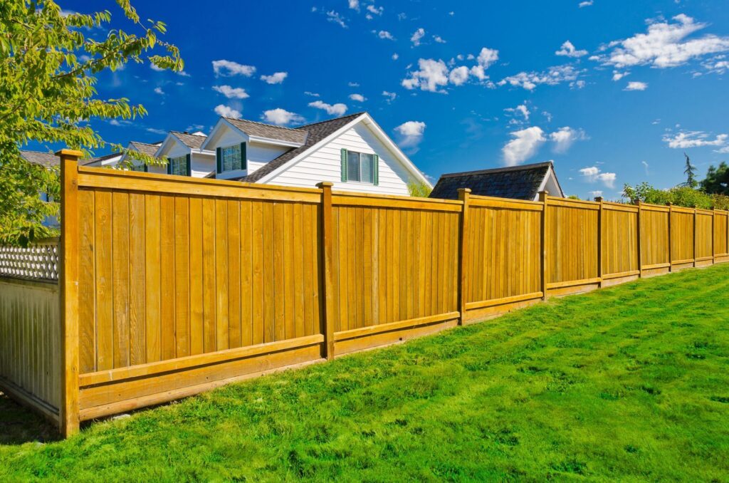 A natural wood privacy fence with capped posts along a sloped green lawn, with white houses and a bright blue sky behind
