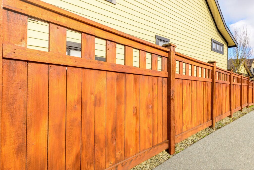 A freshly stained reddish-brown cedar fence with a spindle-topped section running beside a pale yellow house on a gravel path