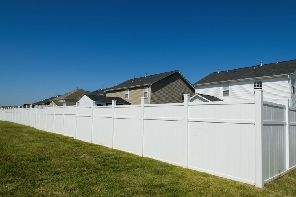 A long white vinyl privacy fence along a grassy yard under a clear blue sky, with two-story houses behind it
