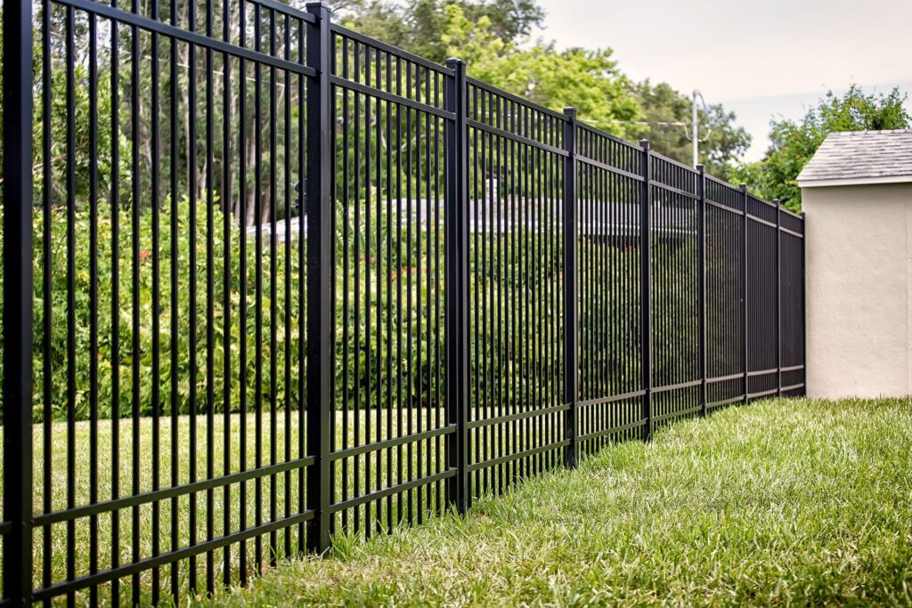 A tall black aluminum picket fence enclosing a landscaped green backyard with shrubs, trees, and a stucco shed