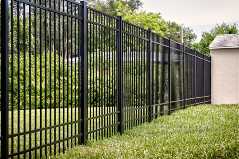 A tall black aluminum picket fence enclosing a landscaped green backyard with shrubs, trees, and a stucco shed