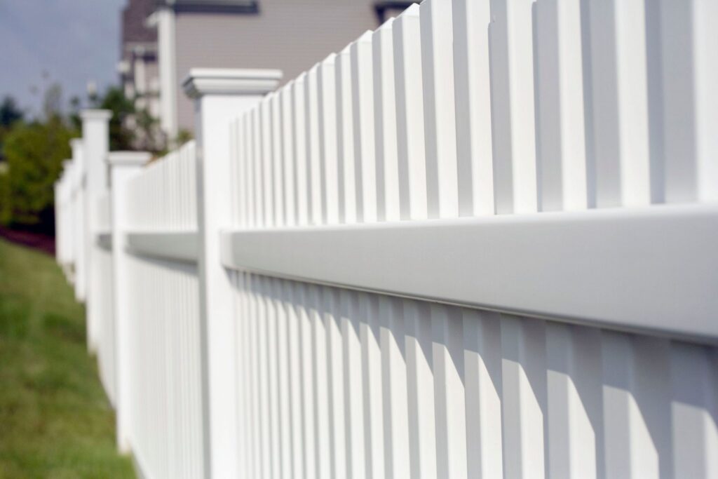 Angled close-up of a white vinyl picket fence with staggered picket tops and decorative post caps beside a green lawn