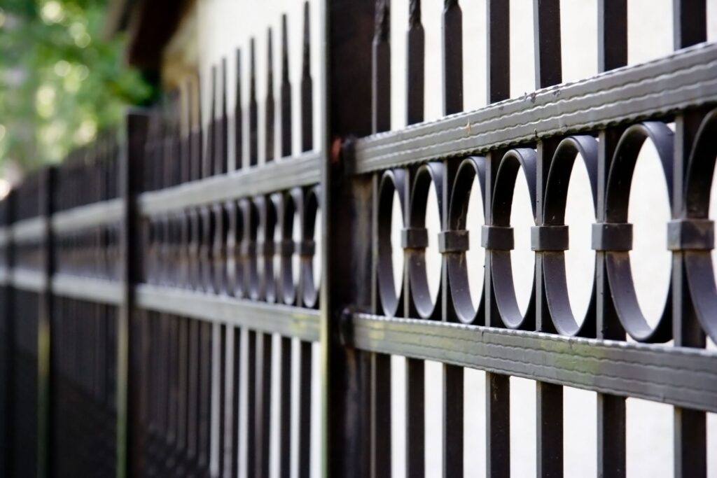 Angled close-up of a black metal fence with decorative circular scroll rings between the pickets, receding toward greenery and a building