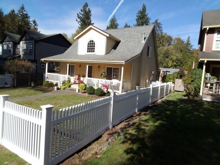A white vinyl scalloped picket fence enclosing the front yard of a yellow two-story cottage with a covered porch, evergreens behind on a sunny day