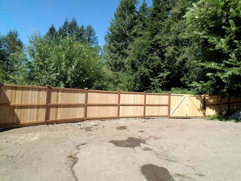 A new cedar privacy fence with a matching double gate running across a bare dirt yard backed by tall evergreen and deciduous trees