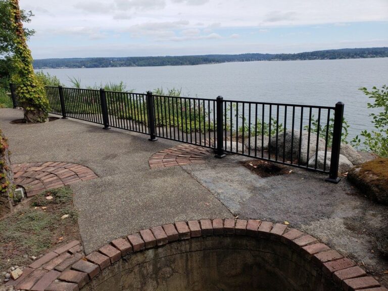 A black aluminum railing along an aggregate-concrete patio with brick accents overlooking a wide bay, with a circular brick fire pit in the foreground