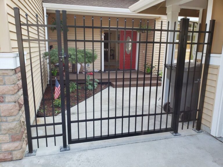 A black aluminum spear-top fence and swing gate enclosing a narrow side yard between a tan house and its walkway