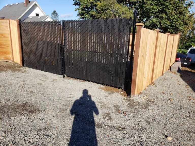 A black chain-link fence with privacy slats and a double gate joining a new cedar privacy fence around a gravel lot, the photographer's shadow in the foreground