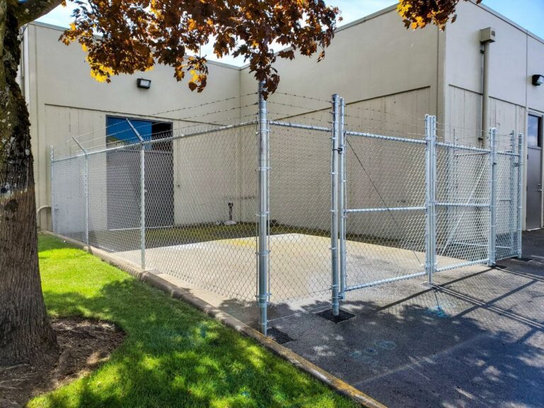 A tall galvanized chain-link enclosure topped with barbed wire wrapping a concrete pad against a large tan commercial building, framed by autumn maple leaves