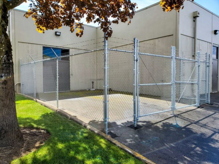 A tall galvanized chain-link enclosure topped with barbed wire against a large tan commercial building, framed by autumn leaves