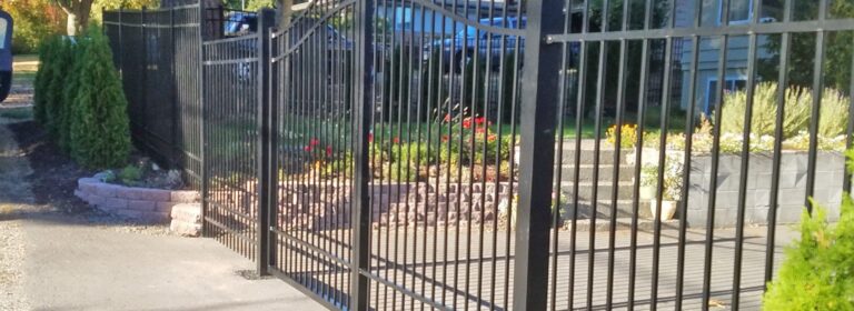 A black aluminum arched driveway gate and matching fence enclosing a front yard with flower beds and a stone retaining wall, a blue house behind in evening light