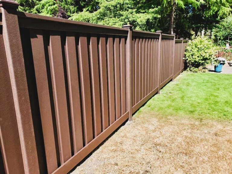 A brown vinyl privacy fence with framed panels and capped posts along a green lawn with patchy dry grass, evergreens behind on a sunny day