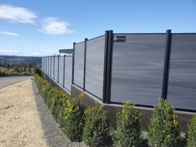 A modern gray composite horizontal-plank privacy fence with black aluminum posts on a concrete base, lined with young boxwood shrubs, overlooking a bay