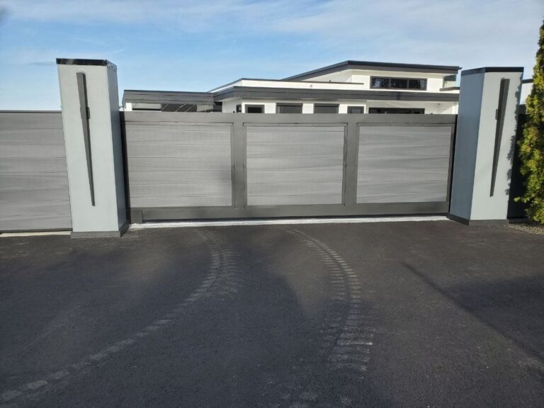 A modern gray composite sliding driveway gate with a black frame between tall light-gray pillars accented with vertical black bars, in front of a contemporary house