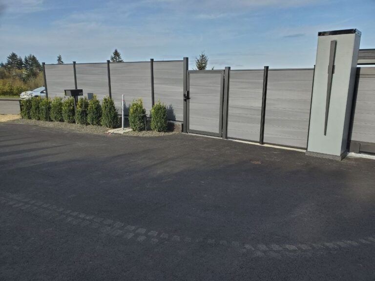 A long gray composite horizontal-plank privacy fence with a matching pedestrian gate and modern pillar, bordered by young boxwood shrubs and a mailbox along a paved driveway