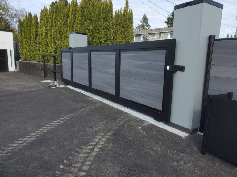 A dark-framed gray composite cantilever sliding gate partly open on its track, with a modern gray pillar and arborvitae hedge behind, on a fresh asphalt drive
