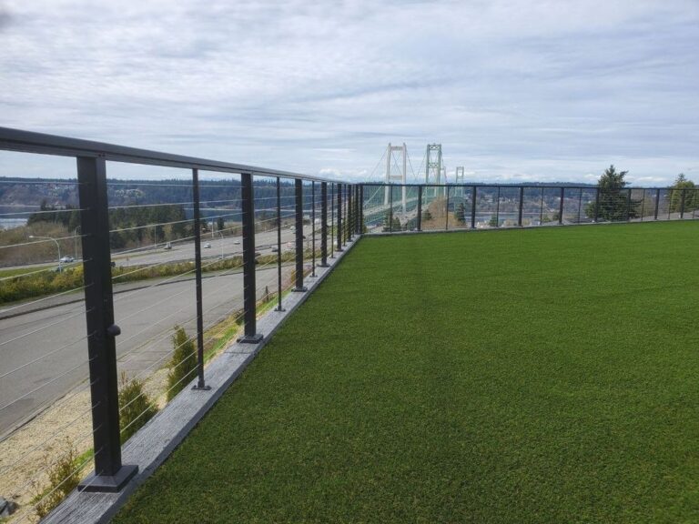 A black horizontal cable railing along the edge of an artificial-turf rooftop lawn, overlooking a highway with the Tacoma Narrows Bridge in the distance