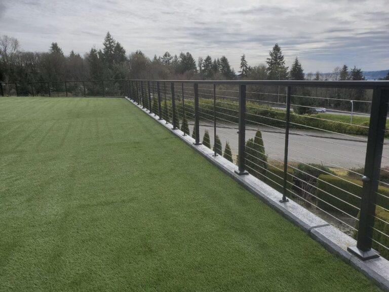 A black metal cable railing bordering a large artificial-turf lawn on a terraced property, overlooking a road and evergreens under an overcast sky