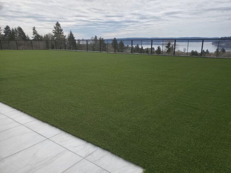 A wide artificial-turf lawn edged with white stone pavers and enclosed by a low black cable railing, with a view of Puget Sound and forested shoreline beyond