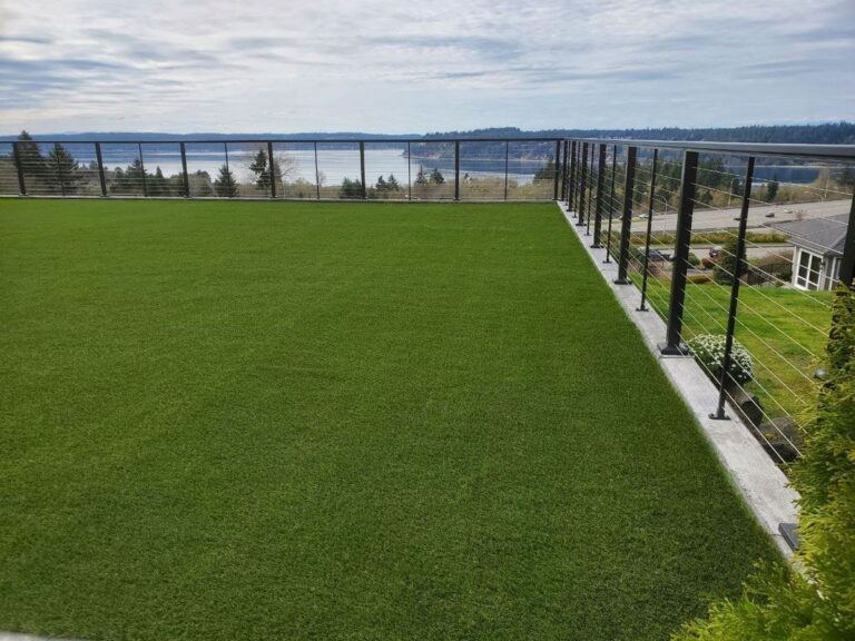 An artificial-turf lawn with a white paver border, ringed by a black metal cable railing overlooking water, hills, and a highway