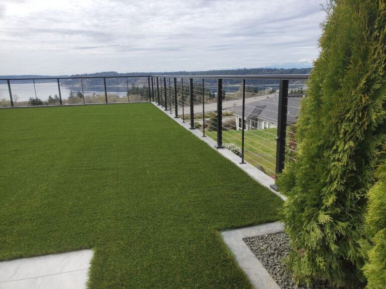 The corner of an artificial-turf lawn with a black cable railing and modern posts, an arborvitae hedge and gravel bed at right, a water view and a house below