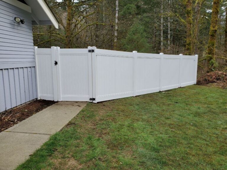 A white vinyl privacy fence with a matching gate running from the side of a gray house across a lawn, bare mossy forest behind on an overcast day