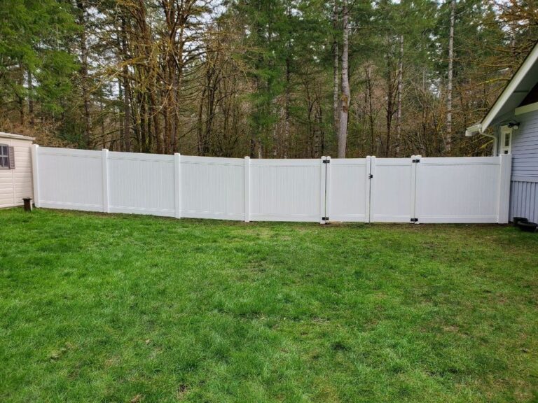 A long white vinyl privacy fence with a double gate enclosing a large grassy backyard beside a gray house, tall leafless trees behind