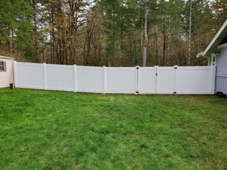 A long white vinyl privacy fence with a double gate enclosing a grassy backyard beside a gray house, bare trees behind