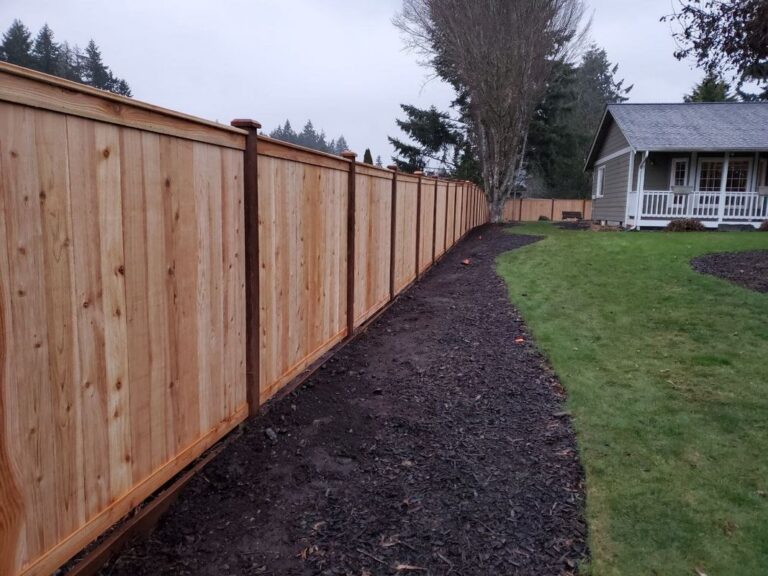 A new golden cedar privacy fence with capped posts running along a fresh dark-mulch bed that curves beside a green lawn toward a gray craftsman house