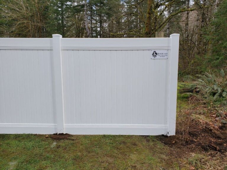 Close-up of a white vinyl privacy fence panel with a small Lionheart Fence Builders placard, on a grassy edge with mossy forest behind