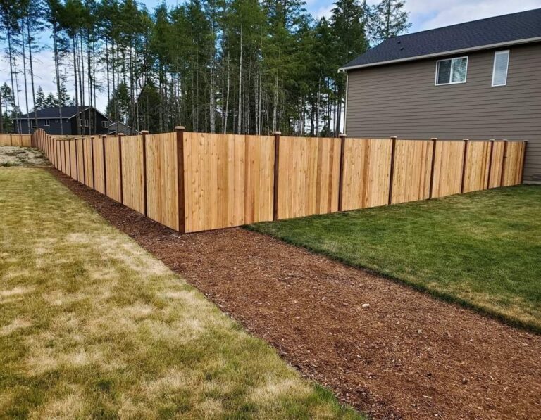 A new unstained cedar privacy fence with capped posts running a long line between a mulch path and lawn, beside a gray two-story house with tall evergreens behind