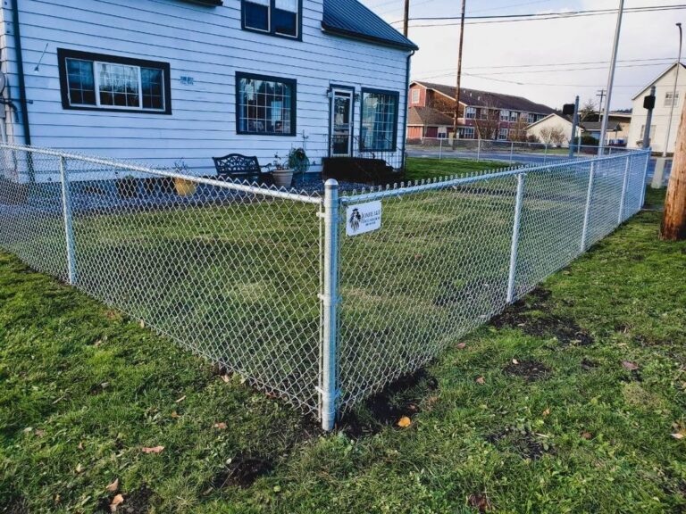 A galvanized chain-link fence with a gate enclosing the side yard of a white two-story farmhouse, with a Lionheart Fence Builders placard on the gate