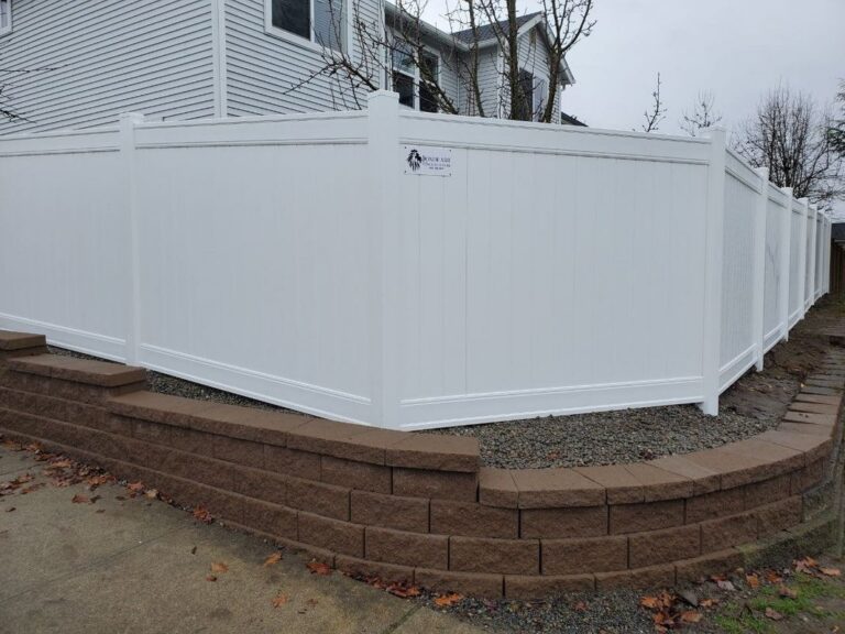 A white vinyl privacy fence wrapping a corner atop a tan retaining-block wall with a gravel bed, in front of a gray two-story house
