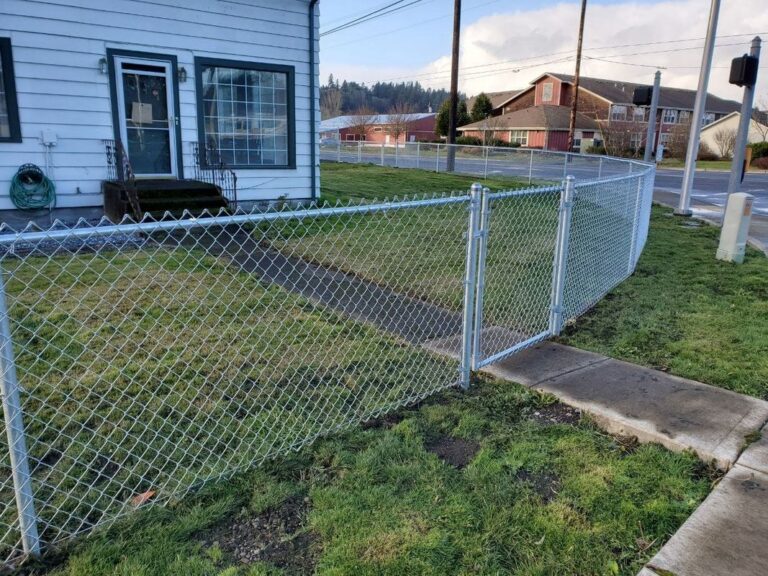 A galvanized chain-link fence with a gate enclosing the side yard of a white house near a street corner, a red barn-style house across the road at dusk