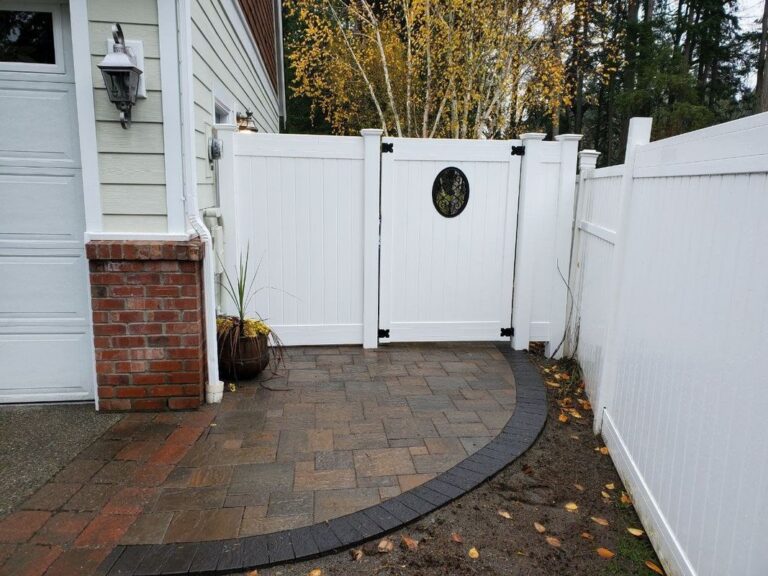 A white vinyl privacy fence with a decorative oval scroll insert in the gate, enclosing a paver walkway beside a house with a brick garage column, autumn birches behind