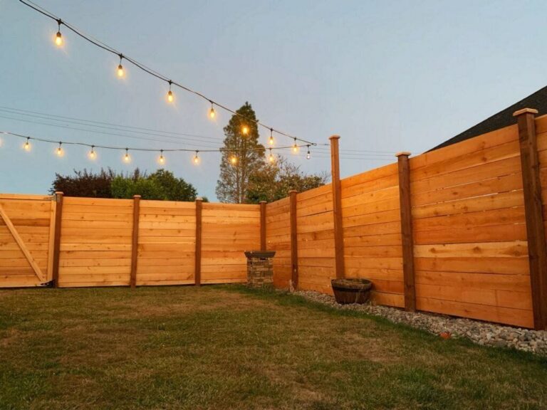 A horizontal-plank cedar privacy fence with capped posts wrapping a backyard at dusk, string lights strung overhead and a stone column along a gravel border