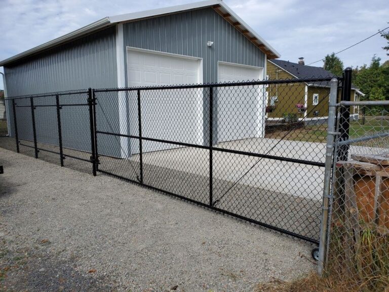 A black chain-link fence with a rolling cantilever gate enclosing the concrete apron of a large gray metal shop building, a yellow house next door