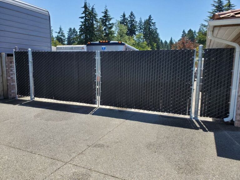 A black chain-link fence with privacy slats and a double swing gate on a concrete driveway screening an RV and trailer, tall evergreens behind on a sunny day