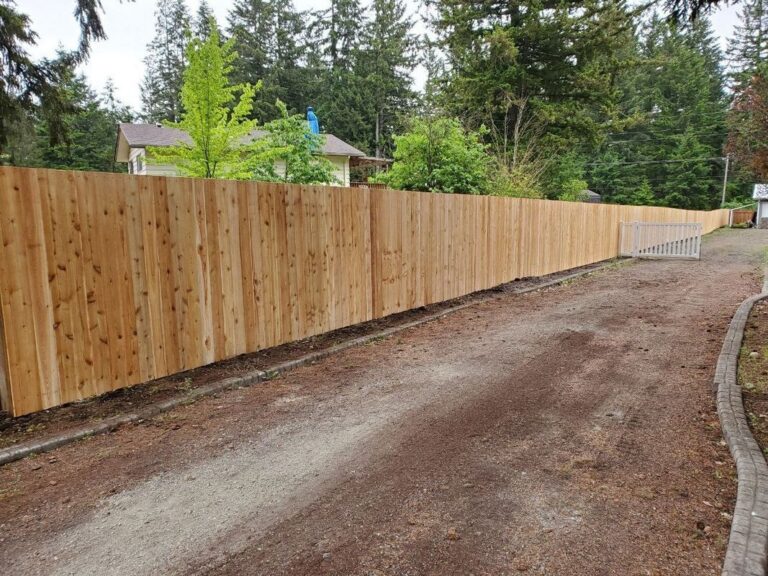 A tall unstained cedar privacy fence running along a gravel driveway toward a small white ranch-style swing gate, forest and a house behind on an overcast day