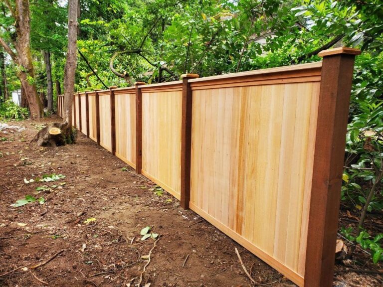 A newly built cedar privacy fence with wide framed panels and capped posts along a cleared, freshly graded yard with green shrubs and trees behind