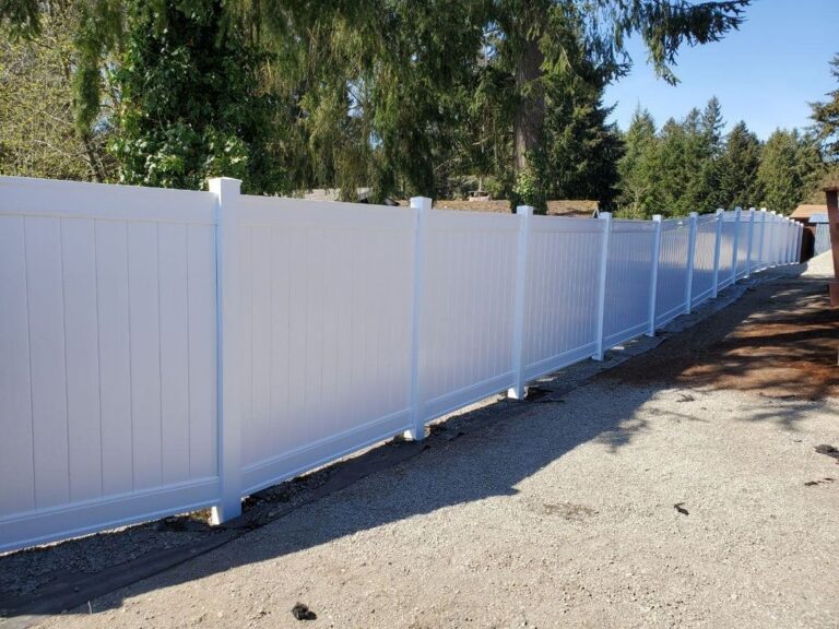 A long white vinyl privacy fence curving along a gravel driveway beside a graded area, with evergreens and blue sky behind