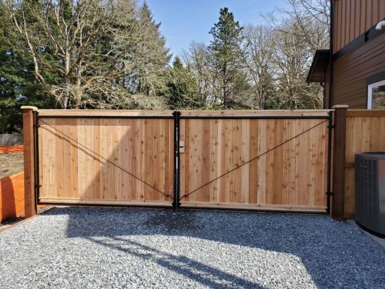 A cedar privacy fence with a black-framed double drive gate with diagonal cable bracing on a gray gravel pad beside a brown two-story house