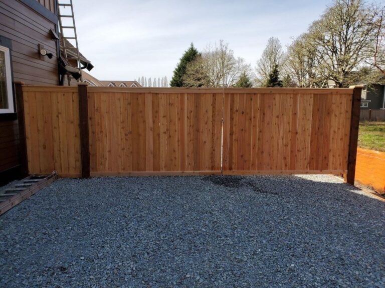 A cedar privacy fence enclosing a fresh gray-gravel side yard beside a dark-sided house, a ladder leaning on the wall, neighborhood and bare trees behind
