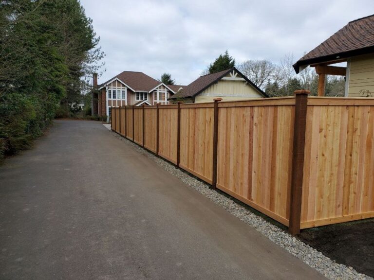 A cedar privacy fence with dark posts running along a paved driveway on a river-rock border, a brick Tudor-style house and a beige house in the background