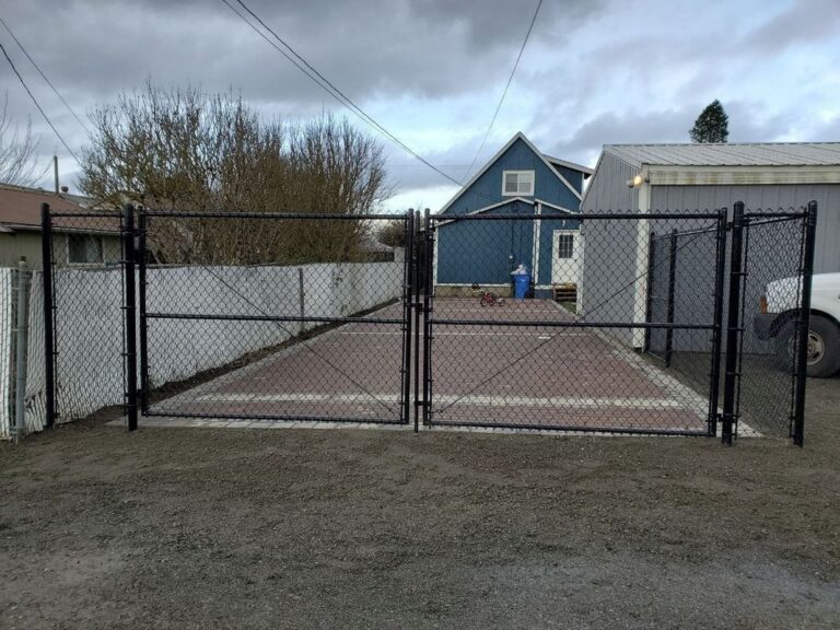 A black chain-link fence with a double swing gate across a paver driveway, enclosing the backyard of a blue house with a metal garage, overcast sky