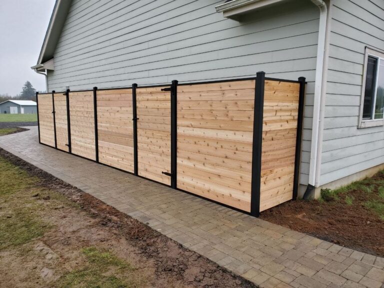 A horizontal-plank cedar privacy fence with black aluminum posts and a gate running along a paver walkway beside a gray house on an overcast day