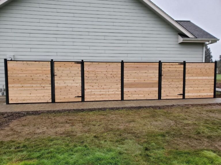 A straight-on view of a horizontal cedar privacy fence with black metal posts and two gates along a paver path beside a gray house
