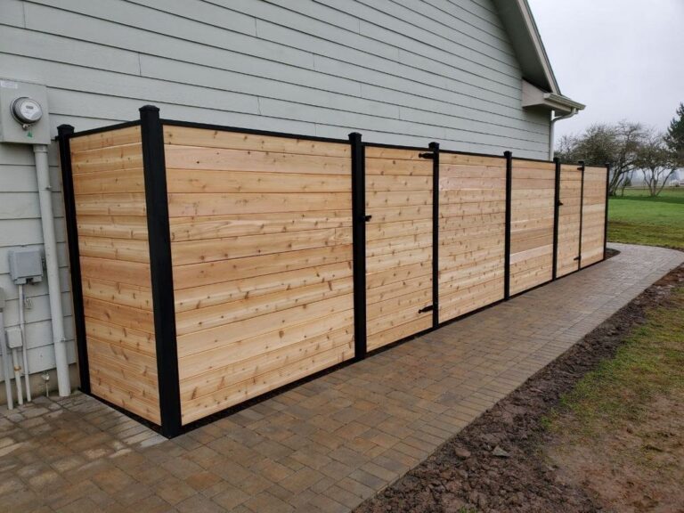 An angled view of a horizontal-plank cedar privacy fence with black posts and gates wrapping the side of a gray house along a paver walkway, an open field beyond