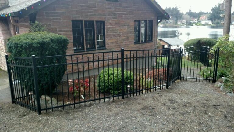 A black aluminum spear-top fence and matching arched gate enclosing a landscaped garden bed in front of a red sandstone lake house, with the lake and homes across the water behind